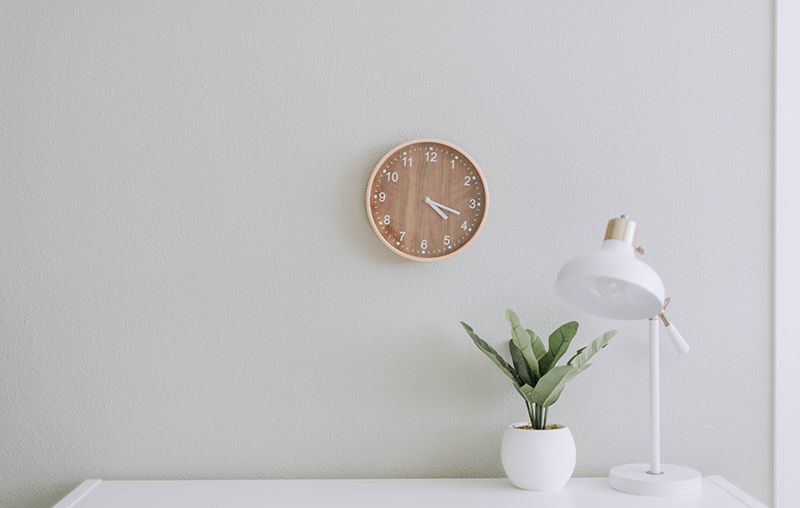A clock mounted on a wall with a table desk underneath holding a plant vase and a lamp.
